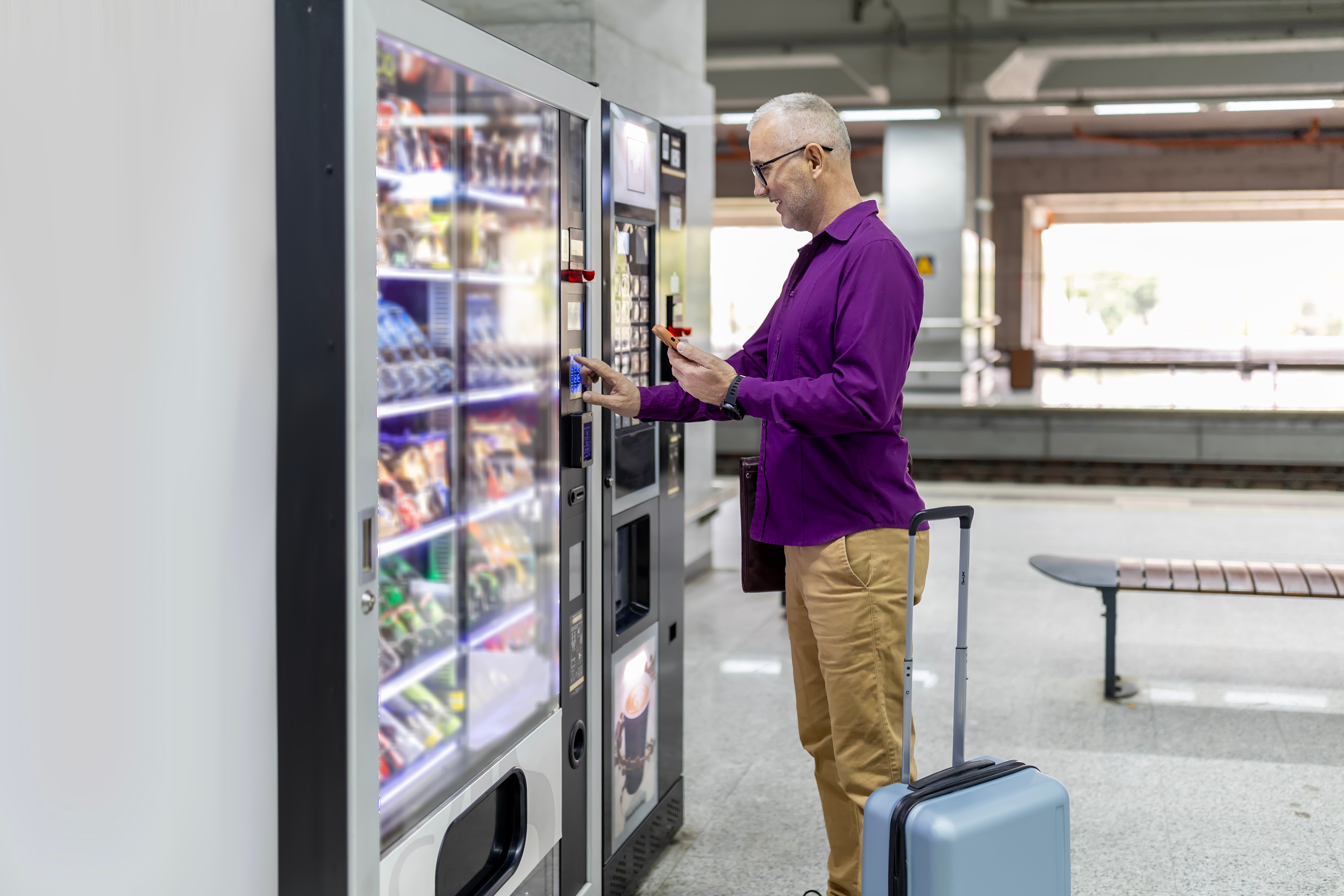 Mature adult man interacting with a modern vending machine using contactless payment via his smartphone, waiting for his train at a station with luggage