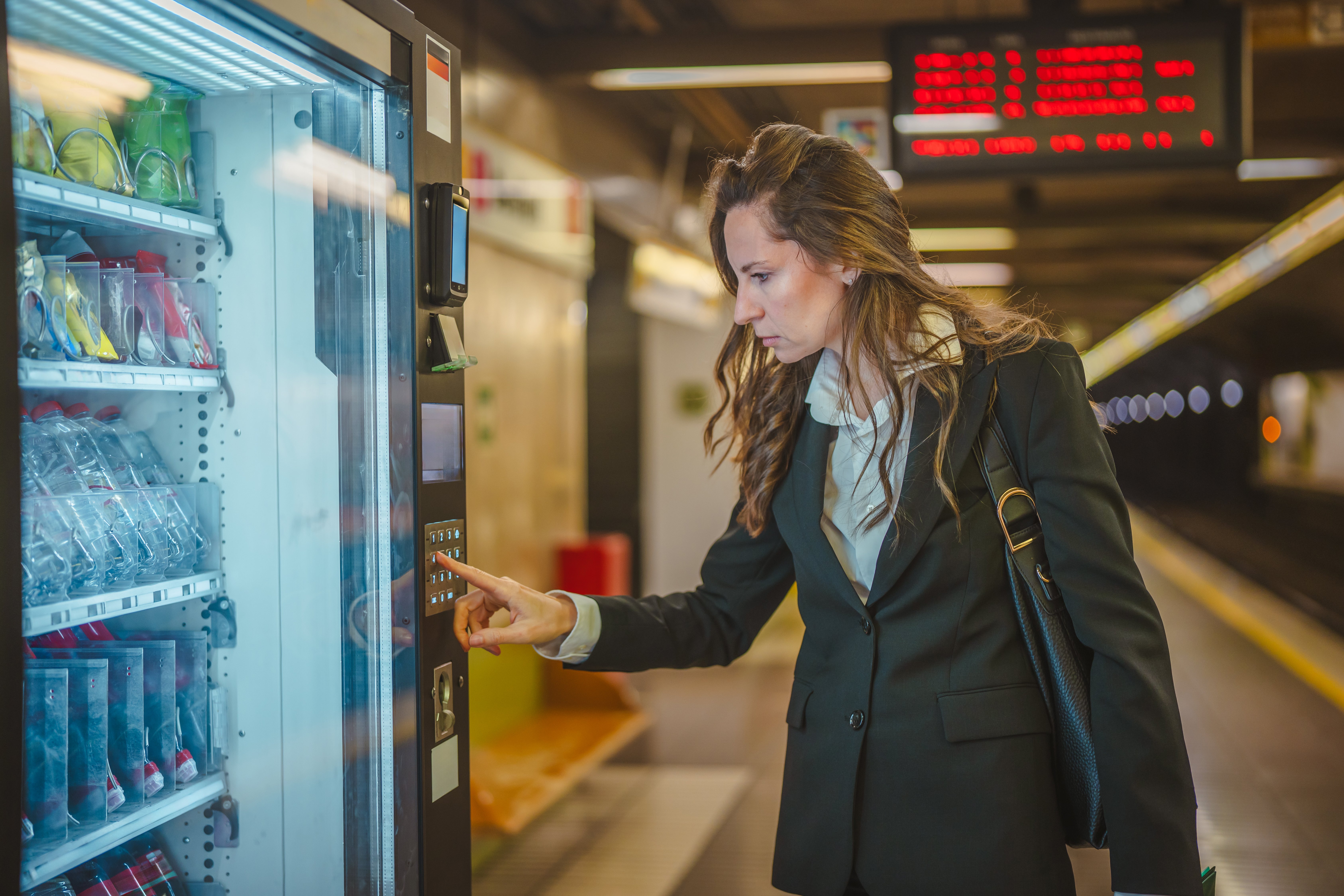 Businesswoman in a suit selecting a snack from an illuminated vending machine, using a keypad to make her purchase while waiting for public transport at a subway platform